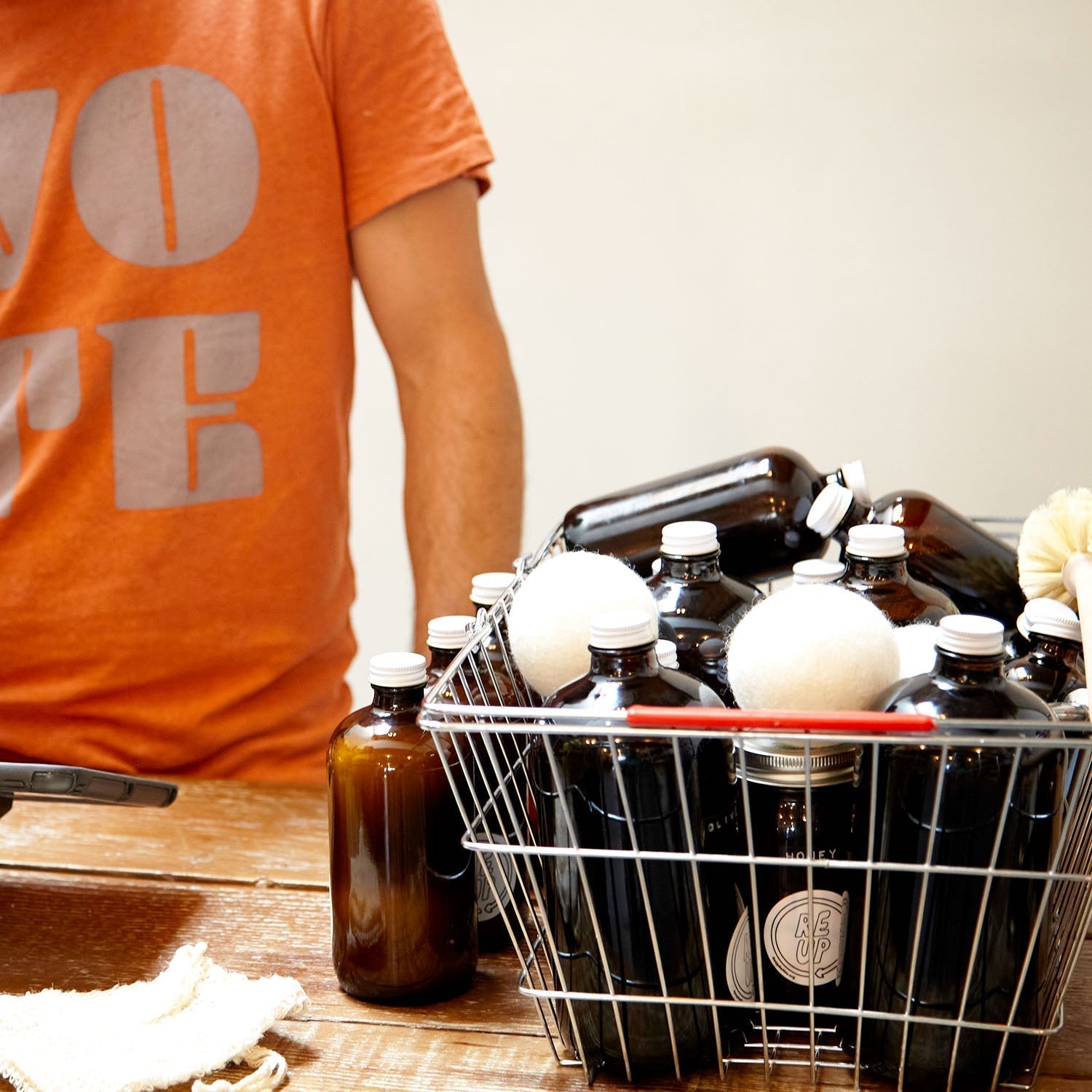 Person standing behind a counter in an orange shirt. On the counter is a basket of refillable amber glass bottles
