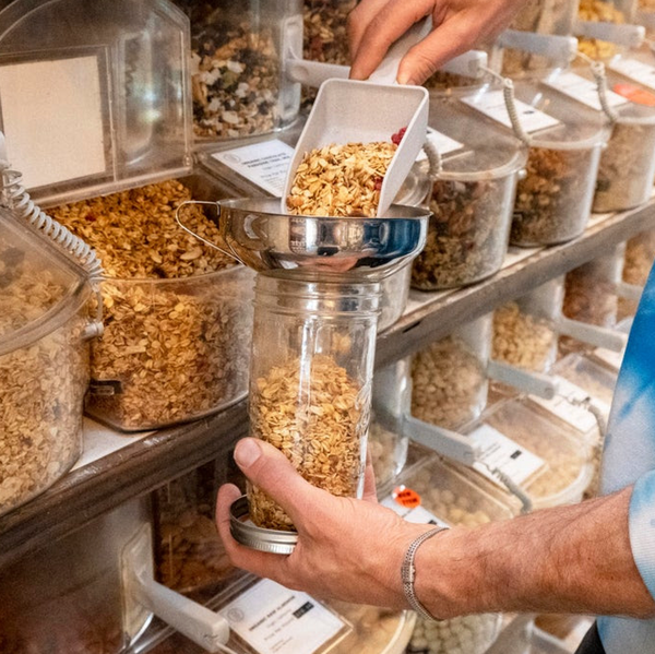 A person wearing a blue tie-dye sweatshirt is scooping granola from a bulk food container into a glass jar using a small white scoop. 